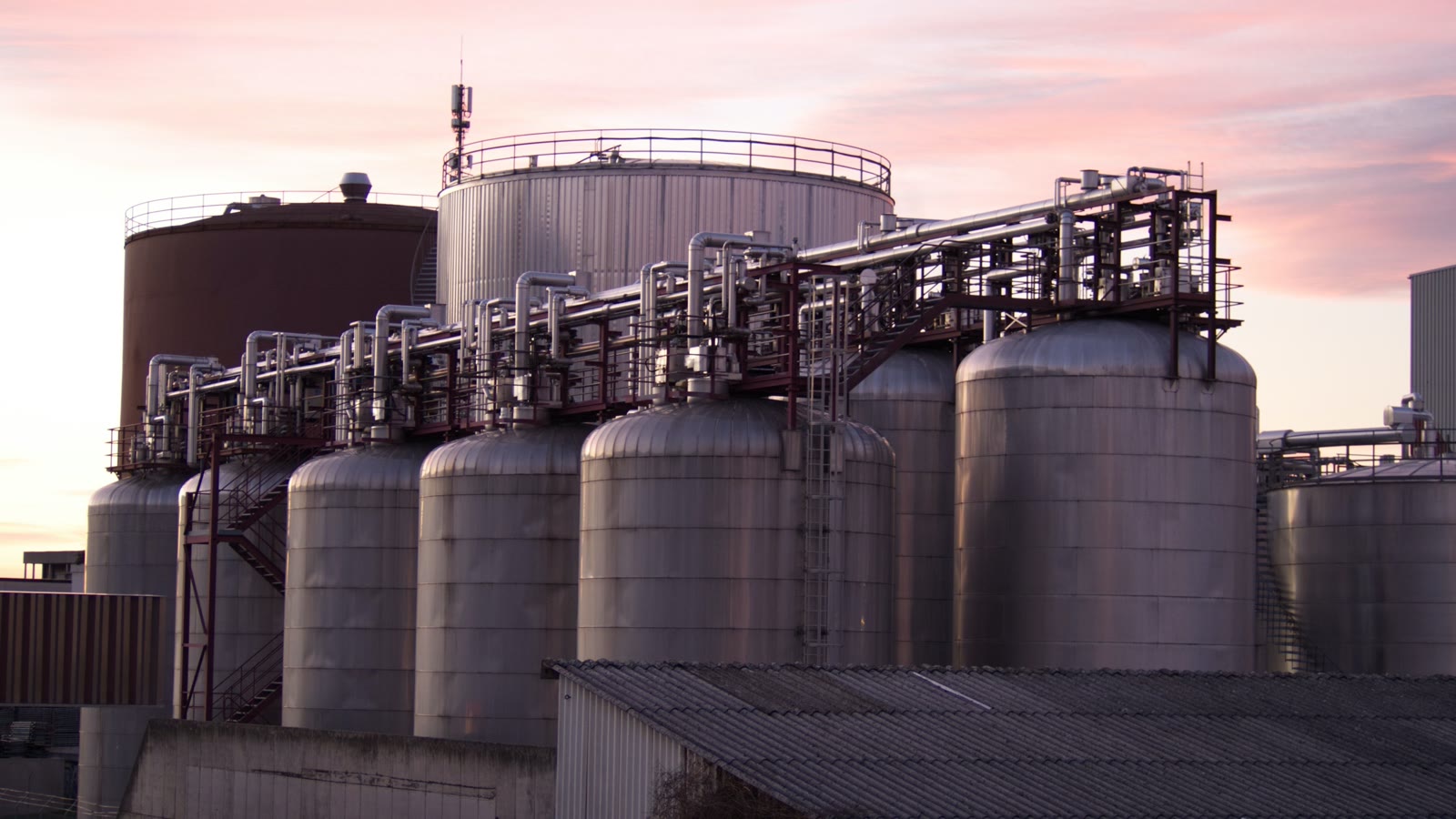 Illustrative refinery storage tanks and pipework at sunset.